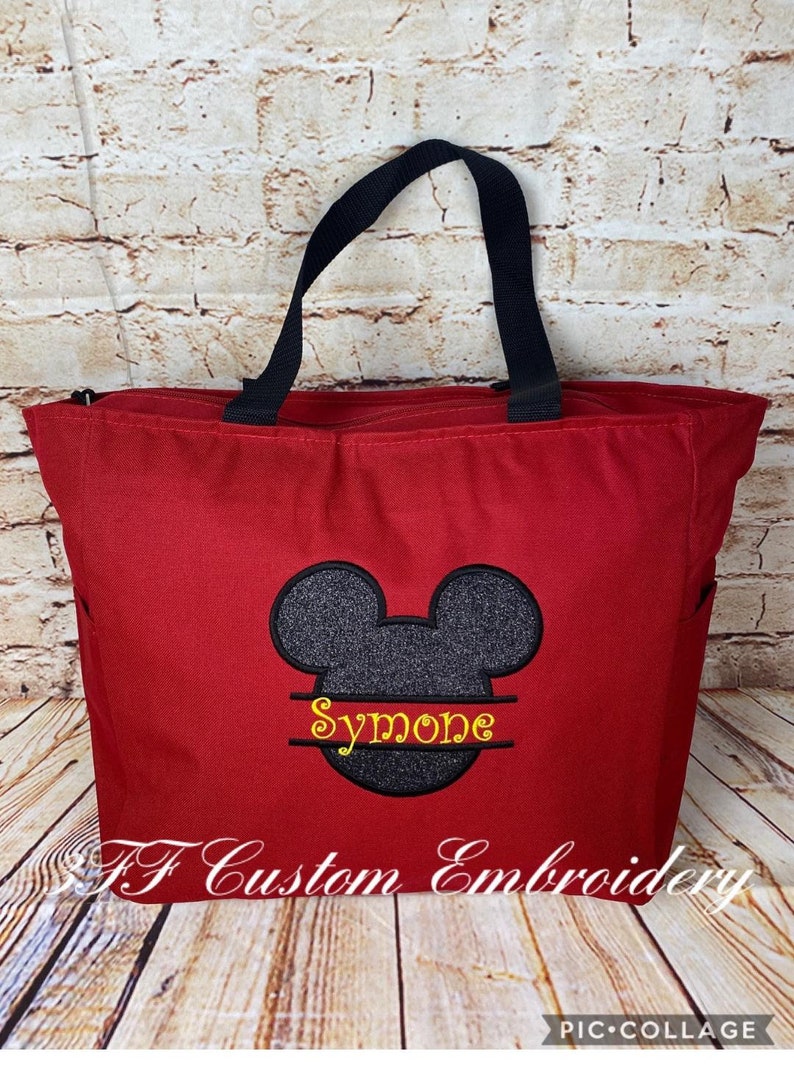 Puede incluir: Bolsa de mano roja con asas negras y una cabeza de Mickey Mouse negra y gris con el nombre "Simone" bordado en amarillo en la parte inferior.