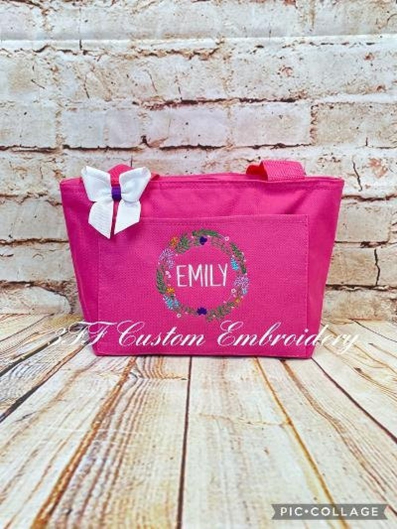 May include: Pink tote bag with a floral wreath design and the name "Emily" embroidered in white. The bag has a white bow on the front.