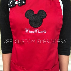 May include: Red apron with a black Mickey Mouse head and the text "Mrs. Martin" embroidered in white. The apron has two pockets and is a custom embroidery product.