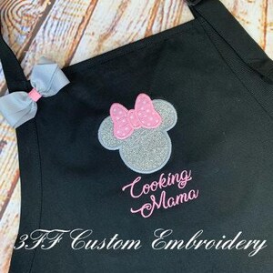 May include: Black apron with a silver and pink embroidered Minnie Mouse head and the text "Cooking Mama". The apron also has the text "3.F Custom Embroidery" at the bottom.