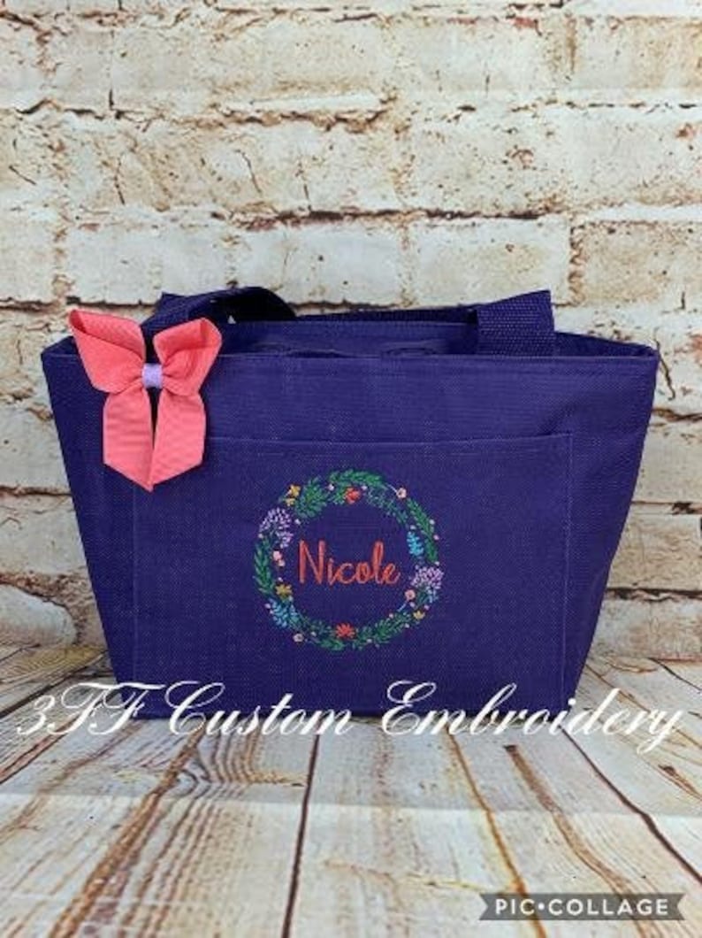 May include: A purple tote bag with a floral wreath design and the name "Nicole" embroidered in the center. A pink bow is attached to the bag.