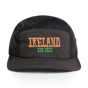 May include: A black five-panel cap with a flat peak. The front panel displays the word "IRELAND" in orange block letters above "EST.1922" in green. The cap is made of a matte material and has a dark grey peak.