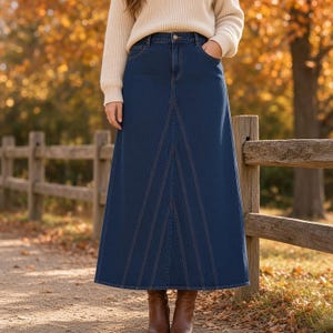 May include: A dark blue denim maxi skirt featuring a high-waisted design with a button and zipper closure, five pockets, and prominent vertical panel stitching that creates a flared silhouette, paired with a cream-colored knit sweater.