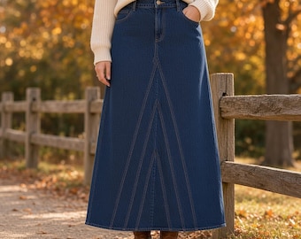 Può includere: Una gonna lunga in denim blu scuro con vita alta, chiusura con bottone e zip, cinque tasche e cuciture verticali in evidenza che creano una silhouette svasata, abbinata a un maglione in maglia color crema.