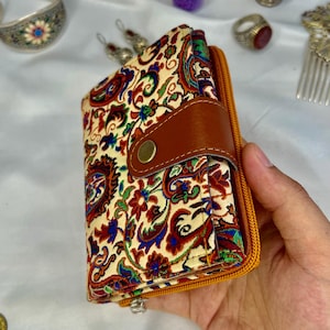 May include: A paisley patterned wallet with a brown leather strap and a gold zip. The wallet is held in a hand and features a button closure. The design includes red, blue, and green floral elements on a cream background.