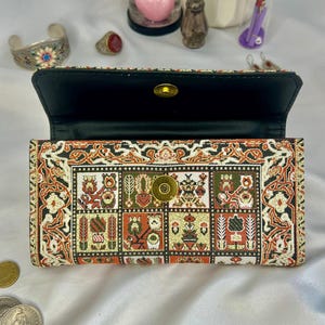 May include: Open wallet with a detailed, colorful pattern. The wallet's exterior features a repeating design in shades of orange, green, black, and cream. The interior is black, and the clasp is gold-toned. A stylish accessory.