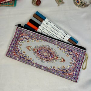 Puede incluir: Un estuche rectangular con un diseño de alfombra tradicional en tonos rojos, azules y dorados. El estuche está abierto, revelando varios rotuladores de colores. Los rotuladores están etiquetados "ART PROFESSIONAL".