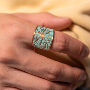 May include: A wide band ring featuring a textured surface with a teal and copper-coloured finish. The design displays an intricate, raised pattern of botanical leaves and stems that wrap around the entire circumference of the metal band.