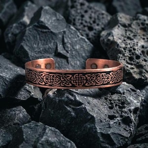 May include: A copper cuff bracelet featuring a continuous black etched Celtic knot pattern around the exterior band. The bracelet has an open design with two small circular magnets visible on the interior ends of the metallic copper band.