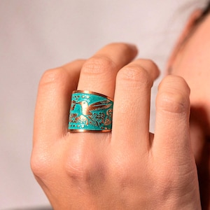May include: A wide copper ring with a turquoise enamel inlay. The ring features a hummingbird design with floral accents. The ring is worn on a finger.
