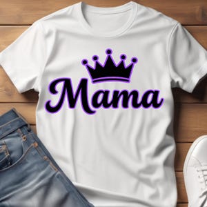 May include: White t-shirt with the word "Mama" in a black script font with a purple outline, topped with a black crown. The shirt is paired with blue jeans and white sneakers, suggesting casual wear.