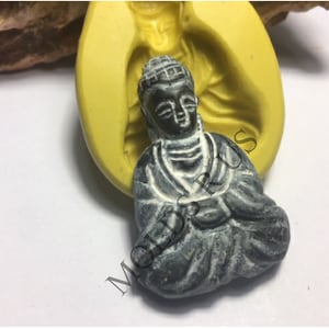May include: A gray resin Buddha statue sitting in a meditative pose with hands clasped in front. The statue is in a yellow silicone mold.