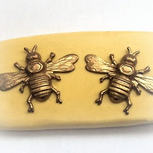 Puede incluir: Un objeto rectangular de color amarillo pálido con dos adornos de abeja de color bronce. Las abejas están detalladas con alas y segmentos corporales. El fondo es de color blanco roto.