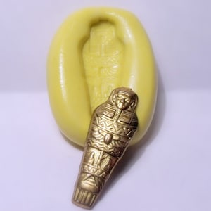 May include: A yellow silicone mold for making a gold-colored resin Egyptian sarcophagus. The mold is shaped like a coffin with a raised design of a pharaoh's head and hieroglyphics.