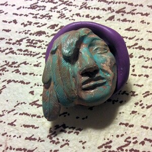 May include: A teal blue and brown clay sculpture of a Native American face with feathers. The sculpture is set against a purple background.