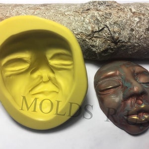 Puede incluir: Un molde de silicona amarillo y una escultura terminada de un rostro humano con los ojos cerrados. La escultura es de color bronce con labios dorados y toques de azul-verdoso. El molde tiene la palabra "MOLDS" impresa.