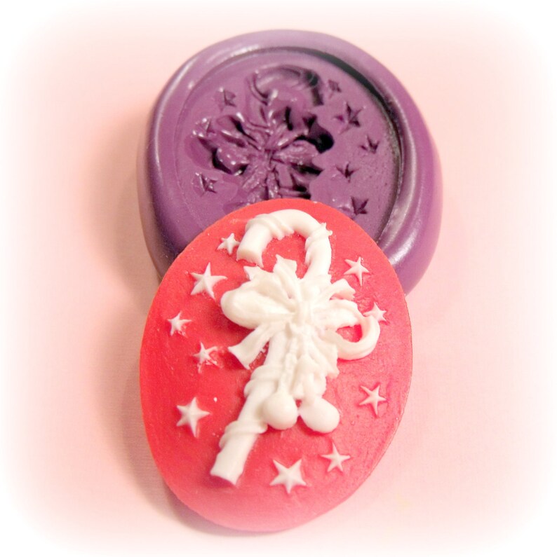 May include: A red silicone mold with a white candy cane design. The mold is shaped like a circle and has a raised design of a candy cane with a bow on top. The candy cane is surrounded by white stars.
