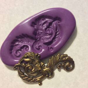 Peut inclure: Un moule en silicone violet avec un design détaillé d'un visage avec des volutes et des feuilles ornées. Un ornement en métal doré assorti avec le même design est présenté à côté du moule.
