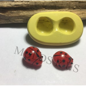 Puede incluir: Un molde de silicona amarillo con dos cavidades, junto a dos figuras de mariquitas rojas. Las mariquitas tienen puntos y marcas negras. El molde y las mariquitas están sobre una superficie blanca.