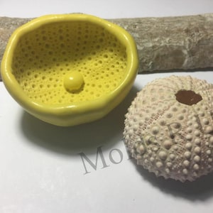 Pode incluir: Uma tigela de cerâmica amarela com uma superfície texturizada e uma pequena depressão redonda no centro. Ao lado da tigela, há um ouriço-do-mar branco com um centro castanho.