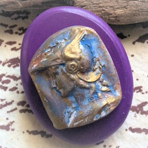 May include: A purple oval-shaped wooden base with a gold and blue ceramic cameo of a winged figure with a helmet. The cameo has a distressed finish.
