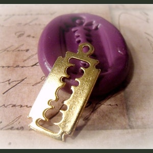 May include: A gold-coloured metal razor blade charm pendant. The charm is hanging from a purple wax seal.