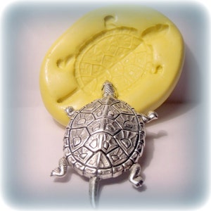 May include: A silver turtle charm with a detailed shell design. The charm is shown next to a yellow silicone mould of a turtle.