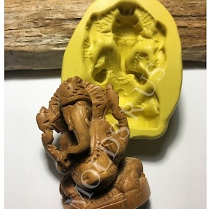 May include: A carved wooden statue of the Hindu god Ganesha, with an elephant head and multiple arms, sits next to a yellow silicone mold. The statue is brown and detailed, while the mold shows the same figure.