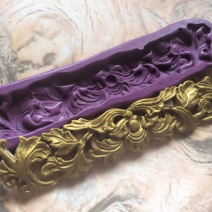 Op de afbeelding: Een paarse siliconen mal met een gedetailleerd bloemenontwerp. De mal wordt gebruikt om een ​​gouden decoratief stuk met een vergelijkbaar bloemenontwerp te maken.