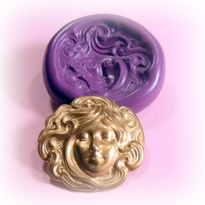 May include: A gold-coloured metal mould of a woman's face with flowing hair. The mould is sitting on top of a purple silicone mould of a similar design.