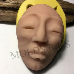May include: A yellow silicone mold of a human face with closed eyes and a relaxed expression. The mold is on a wooden surface. MOLDS R US is printed on the surface.