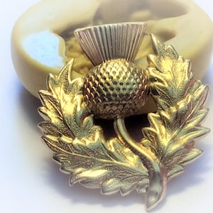 May include: A gold-colored metal thistle with a detailed design, surrounded by a wreath of leaves. The thistle is a traditional symbol of Scotland.