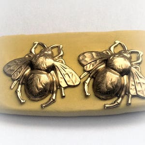 Puede incluir: Dos abalorios de abeja de color dorado con alas y cuerpos detallados. Los abalorios están sobre una superficie de color marrón claro.