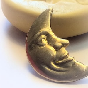 Op de afbeelding: Een gouden halve maan hanger met een gezicht dat in het oppervlak is gebeeldhouwd. Het gezicht heeft een gesloten oog, een neus en een glimlachende mond.