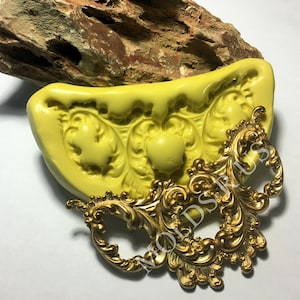 May include: A yellow silicone mold with a design of three ornate floral shapes. A gold metal decorative piece with a similar design is shown next to the mold.