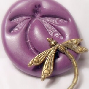 May include: A purple silicone mold with a dragonfly design. A gold-colored dragonfly is sitting on the mold.