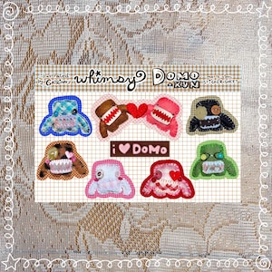 May include: A matte vinyl sticker sheet featuring whimsical Domo-Kun illustrations. The sheet includes various Domo-Kun designs in blue, brown, pink, green, and black, with heart and tooth details. Text includes "whimsy Domo-Kun" and "I ❤️ Domo".