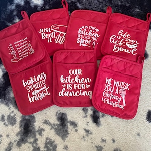 May include: A selection of red quilted oven mitts, each with a different white slogan and graphic. Phrases include "Our Kitchen is for Dancing" and "Life is Short, Lick the Bowl." Each mitt has a pocket and a hanging loop.