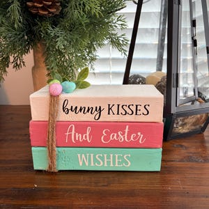 May include: A stack of painted wooden blocks in pastel shades, featuring the phrases "bunny KISSES", "And Easter", and "WISHES". The blocks are tied with twine and adorned with artificial Easter eggs and foliage.