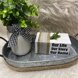 Puede incluir: Una bandeja decorativa de metal con un jarrón blanco, un lazo a cuadros blanco y negro y una pequeña planta verde. La bandeja también contiene tres libros blancos con las palabras "Our Life, Our Story, Our Home" y una cinta de arpillera y encaje.
