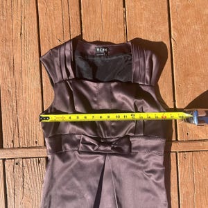 K&ouml;nnte beinhalten: &Auml;rmelloses Satinkleid in einem ged&auml;mpften Lila-Braun-Ton. Das Kleid hat eine Schleife an der Taille und Falten am Oberteil. Das Kleid ist etwa 43 cm breit.