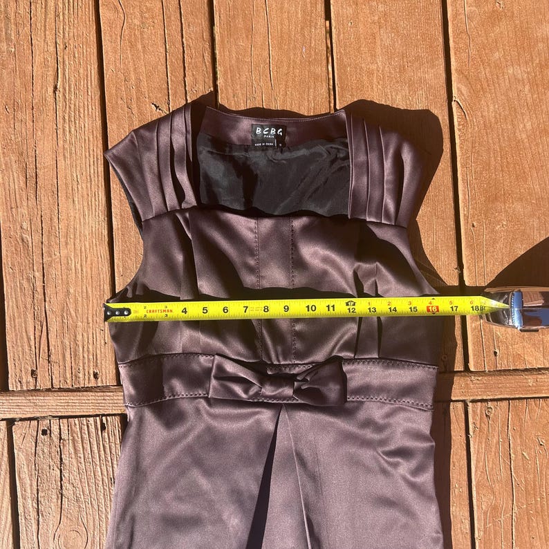K&ouml;nnte beinhalten: &Auml;rmelloses Satinkleid in einem ged&auml;mpften Lila-Braun-Ton. Das Kleid hat eine Schleife an der Taille und Falten am Oberteil. Das Kleid ist etwa 43 cm breit.