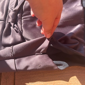 K&ouml;nnte beinhalten: Nahaufnahme eines dunkelvioletten Satin-Kleidungsst&uuml;cks mit Schleifendetail. Der Stoff hat einen subtilen Glanz, und die N&auml;hte sind sichtbar. Eine Hand ber&uuml;hrt den Stoffrand.