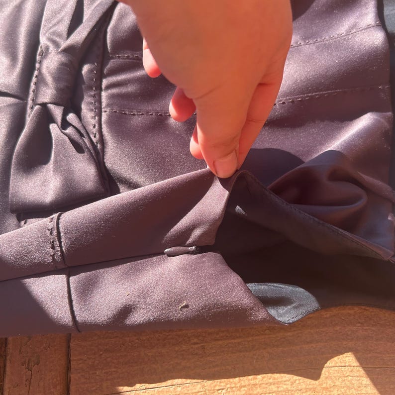 K&ouml;nnte beinhalten: Nahaufnahme eines dunkelvioletten Satin-Kleidungsst&uuml;cks mit Schleifendetail. Der Stoff hat einen subtilen Glanz, und die N&auml;hte sind sichtbar. Eine Hand ber&uuml;hrt den Stoffrand.