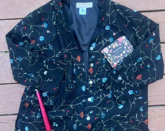 May include: A black long-sleeve button-down shirt featuring an embroidered pattern of swirling vines and colorful flowers in red, blue, and green. The shirt has a collar, white buttons, and a label reading "Norm Thompson". A book titled "Cranford" is placed on the front.