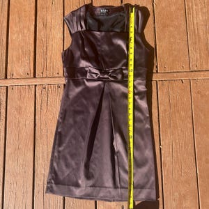 K&ouml;nnte beinhalten: Ein &auml;rmelloses, dunkelbraunes Satinkleid mit Schleife in der Taille. Das Kleid hat einen Faltenrock und einen quadratischen Ausschnitt. Das Kleid ist etwa 61 cm lang.