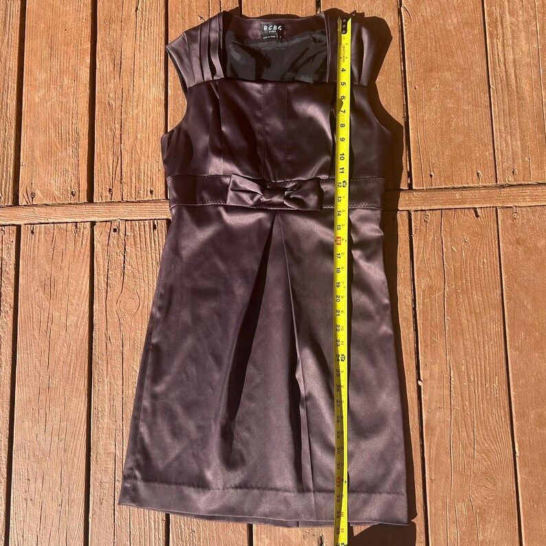 K&ouml;nnte beinhalten: Ein &auml;rmelloses, dunkelbraunes Satinkleid mit Schleife in der Taille. Das Kleid hat einen Faltenrock und einen quadratischen Ausschnitt. Das Kleid ist etwa 61 cm lang.