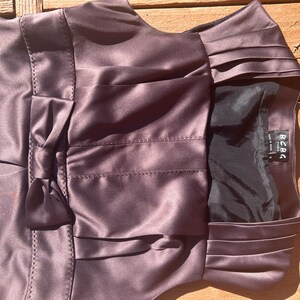 K&ouml;nnte beinhalten: Ein pflaumenfarbenes Satinkleid mit Schleife und Falten. Das Kleid hat ein figurbetontes Oberteil und einen ausgestellten Rock. Das Etikett "BCBG Paris" ist sichtbar. Das Kleid besteht aus einem gl&auml;nzenden, glatten Stoff.