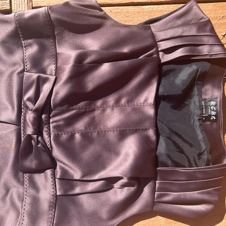 K&ouml;nnte beinhalten: Ein pflaumenfarbenes Satinkleid mit Schleife und Falten. Das Kleid hat ein figurbetontes Oberteil und einen ausgestellten Rock. Das Etikett "BCBG Paris" ist sichtbar. Das Kleid besteht aus einem gl&auml;nzenden, glatten Stoff.