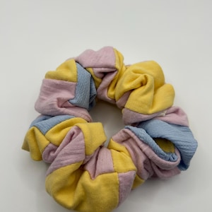May include: A colorful scrunchie with a patchwork design. The fabric is divided into sections of yellow, light blue, and pink. The scrunchie is made of soft fabric and is designed to hold hair in place.
